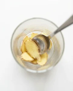 Overhead view of ginger slices being pressed with a spoon in a clear glass on a white surface