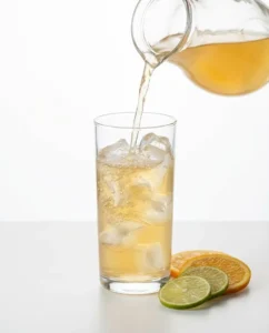 Citrus ginger kombucha being poured into a tall ice filled glass on a white surface