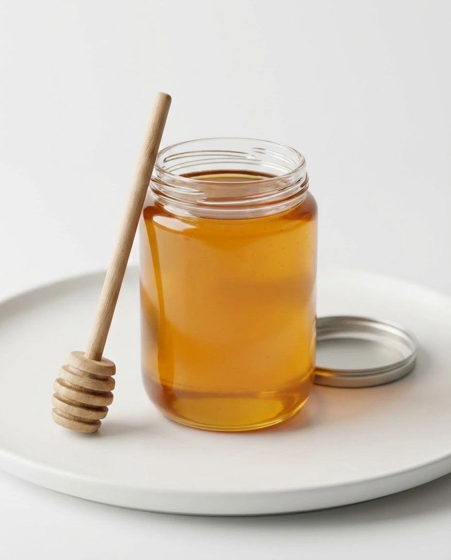 Clear jar of golden honey with a wooden dipper on a clean white background.
