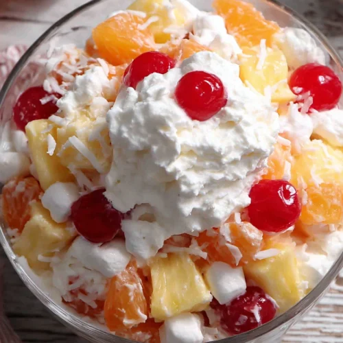 Ambrosia salad with Cool Whip in a glass bowl, featuring pineapple, oranges, marshmallows, coconut, and cherries