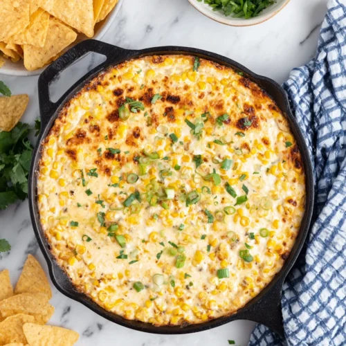 Creamy baked elote dip in a skillet, topped with green onions and cilantro, served with tortilla chips
