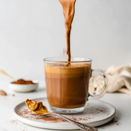 Creamy coffee smoothie being poured into a glass mug with peanut butter and cocoa powder nearby