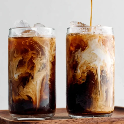 Two glasses of cold brew coffee with cream and ice, swirling together on a wooden tray