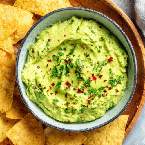 Creamy guasacaca in a white bowl with tortilla chips, garnished with herbs and chili flakes