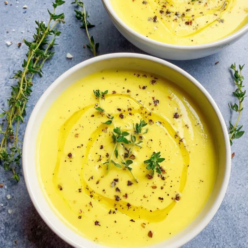Creamy yellow squash soup in a white bowl with olive oil drizzle and fresh herbs