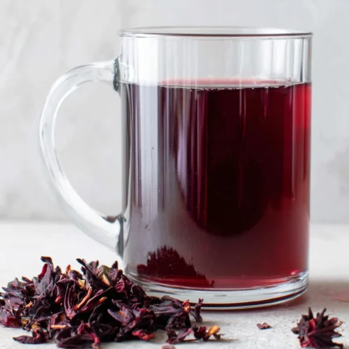A glass mug of vibrant red hibiscus tea with dried hibiscus petals nearby