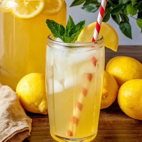 Fresh homemade lemonade in a mason jar with ice, lemon slice, mint, and red striped straw, pitcher in background