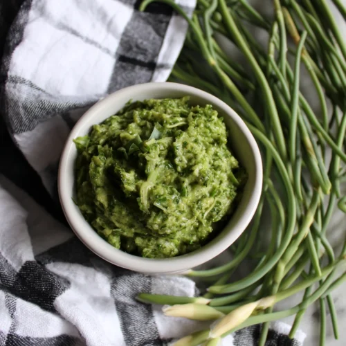 Fresh garlic scape pesto in a white bowl with raw garlic scapes on a checkered cloth