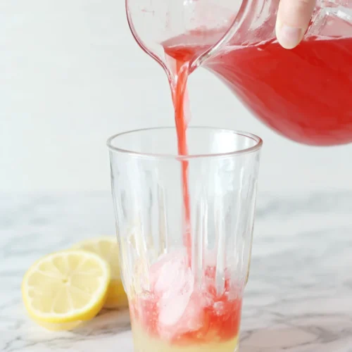 Hand pouring cranberry juice into pink lemonade pitcher with lemon garnish