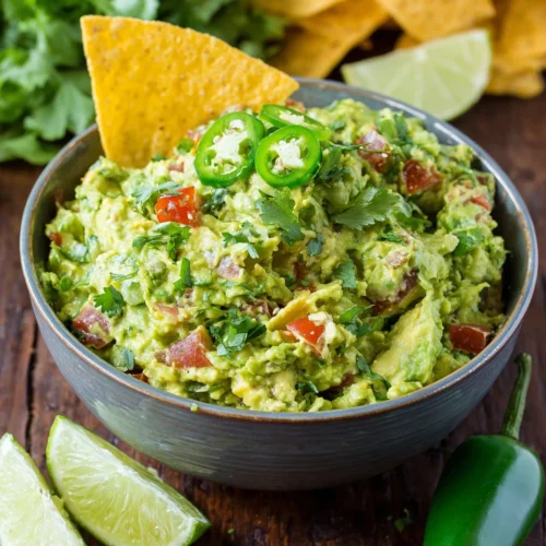 Spicy guacamole in a white bowl with jalapeños and tortilla chip, fresh ingredients visible