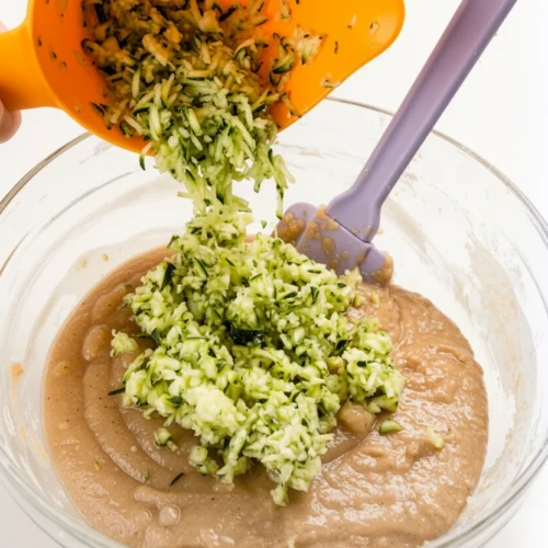 Grated zucchini being added to applesauce zucchini bread batter in a glass bowl